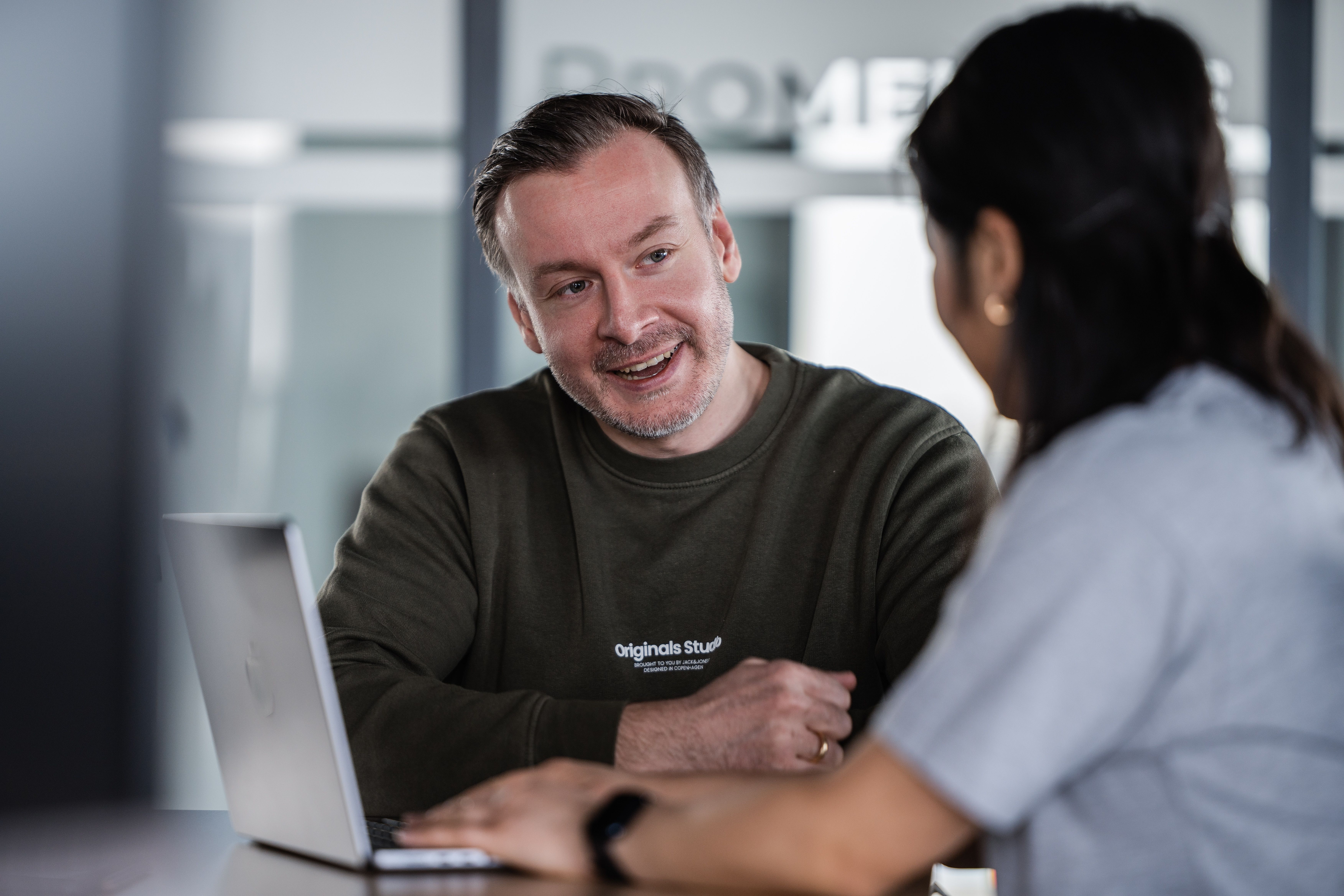 Zwei Personen vor einem MacBook, einer im olivgrünen Pullover lacht der anderen Person zu